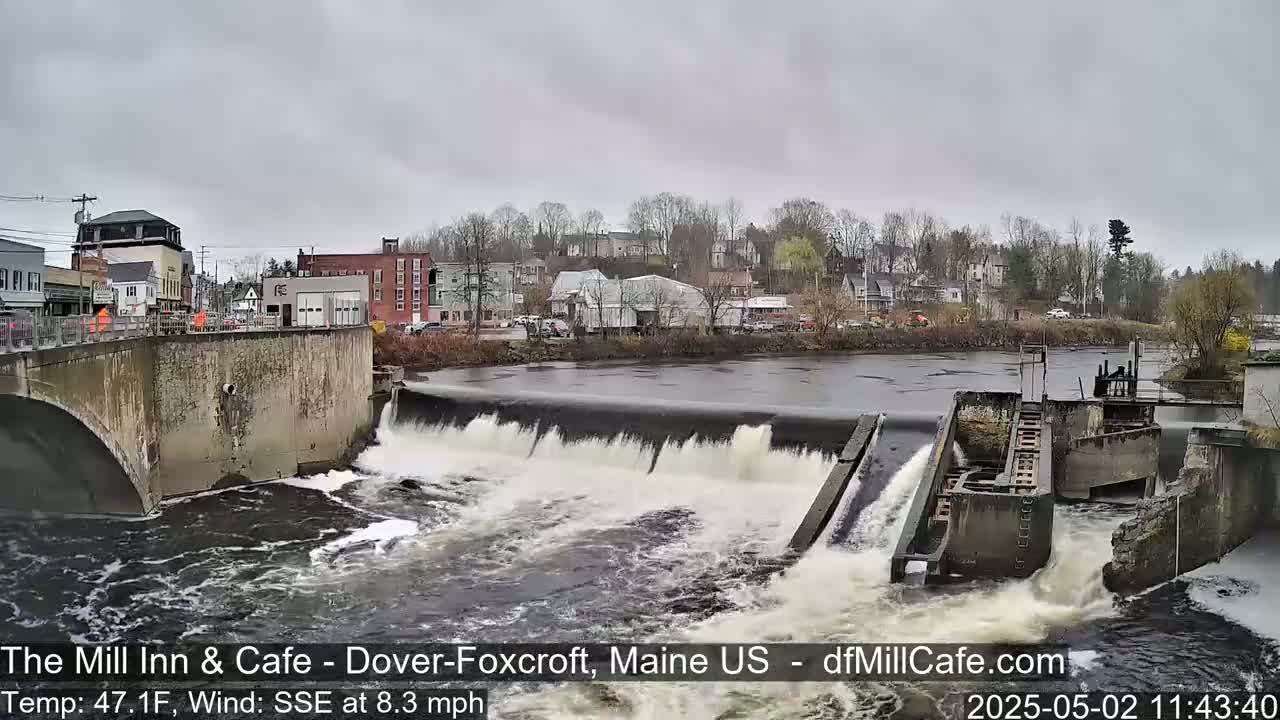 A river flows over a dam in a town under an overcast sky.