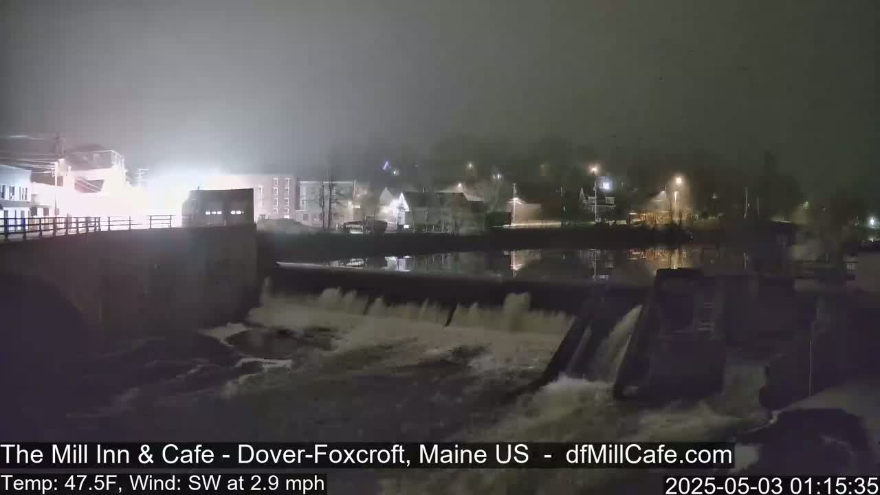 A foggy night scene shows a river flowing over a dam, with buildings and lights visible in the background.