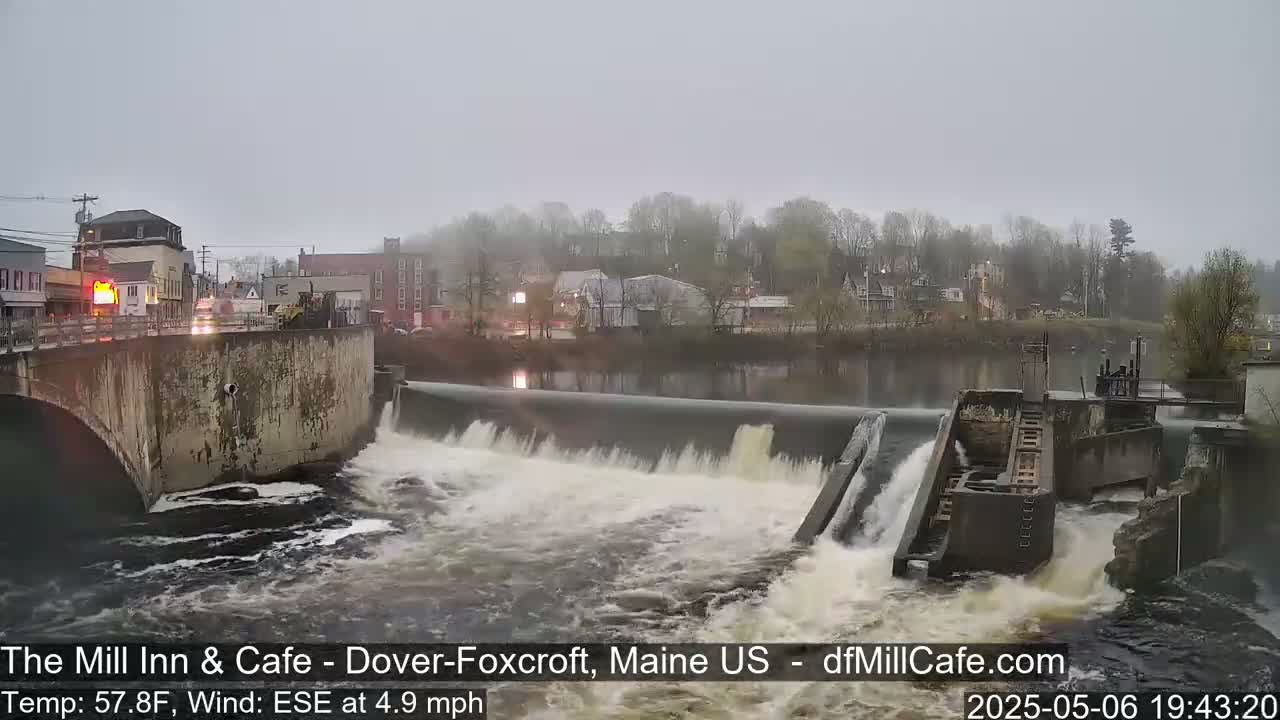 A dam with water flowing over it is situated in a town on an overcast day.