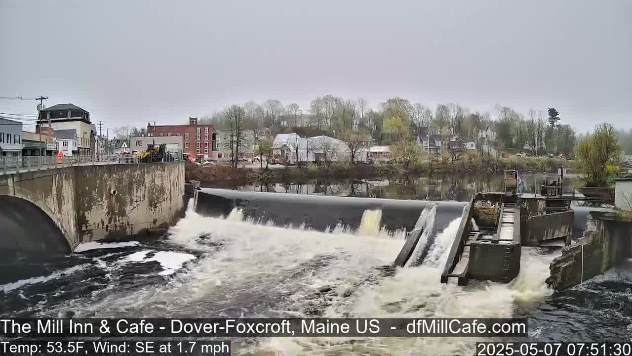 Water rushes over a dam in a town on an overcast day.