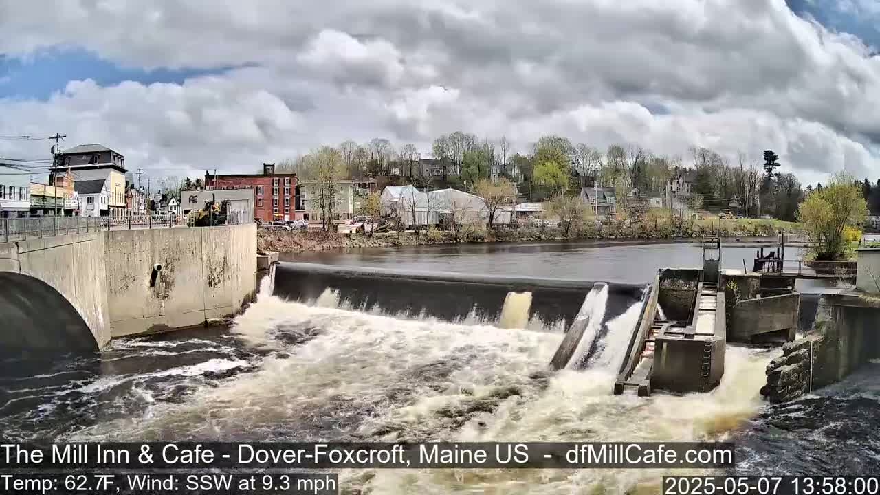 A river flows over a dam in a town, under a partly cloudy sky.