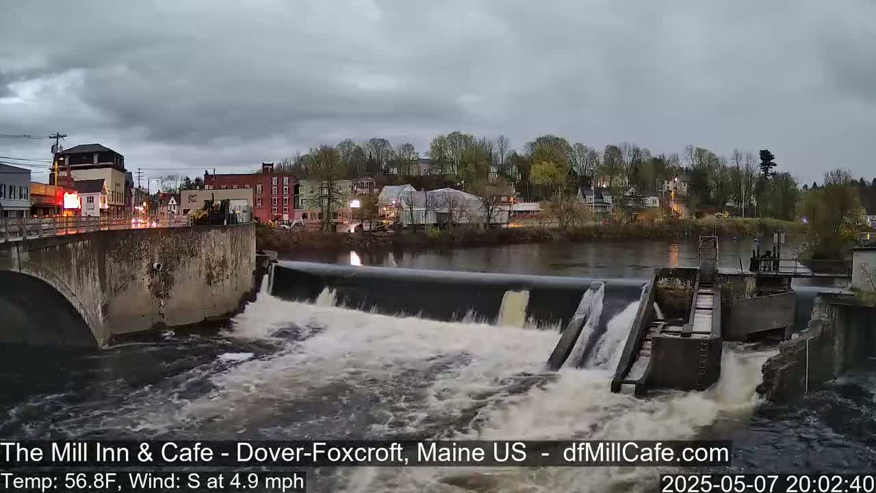 A river flows over a dam in front of a town under an overcast sky.