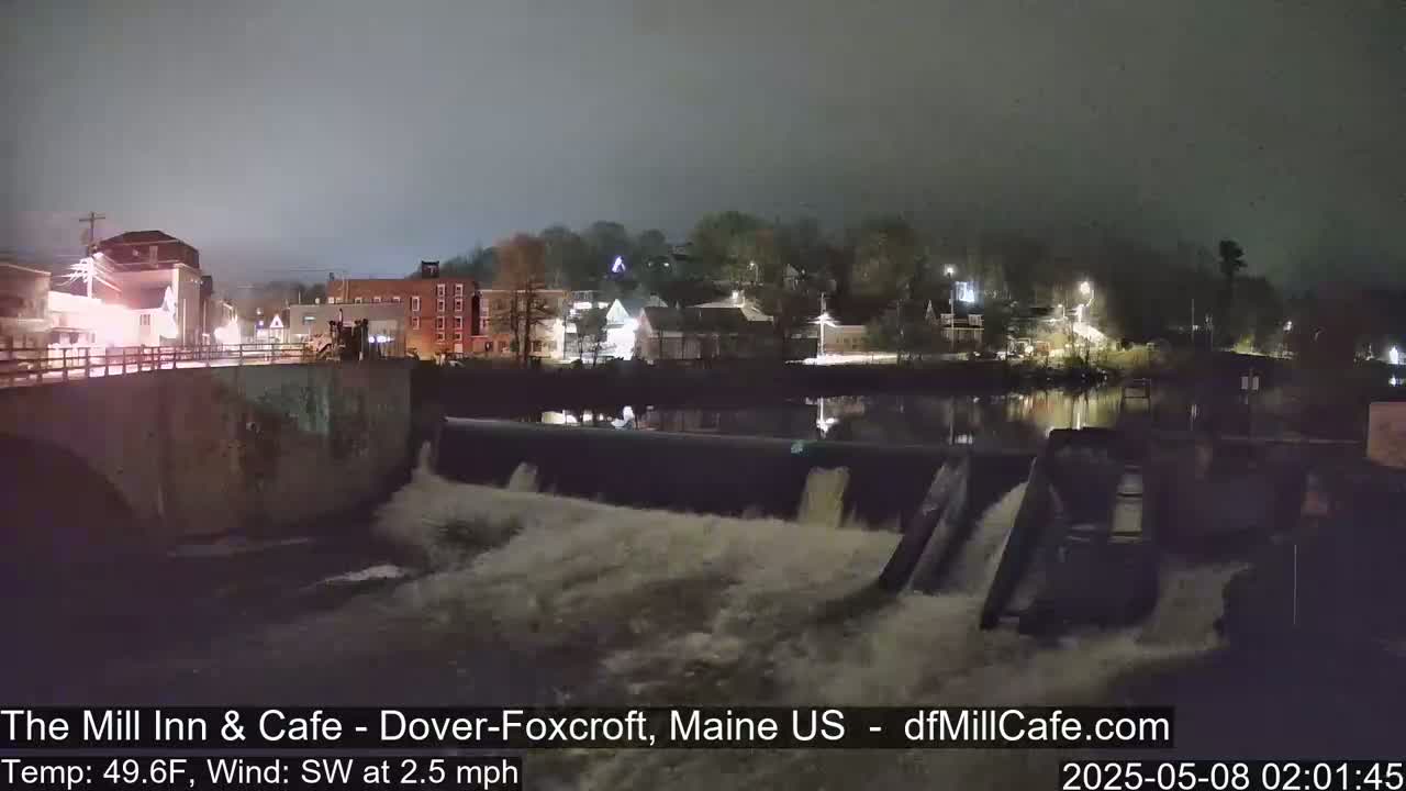 A nighttime view of a town's river dam with water flowing over it under a hazy sky.