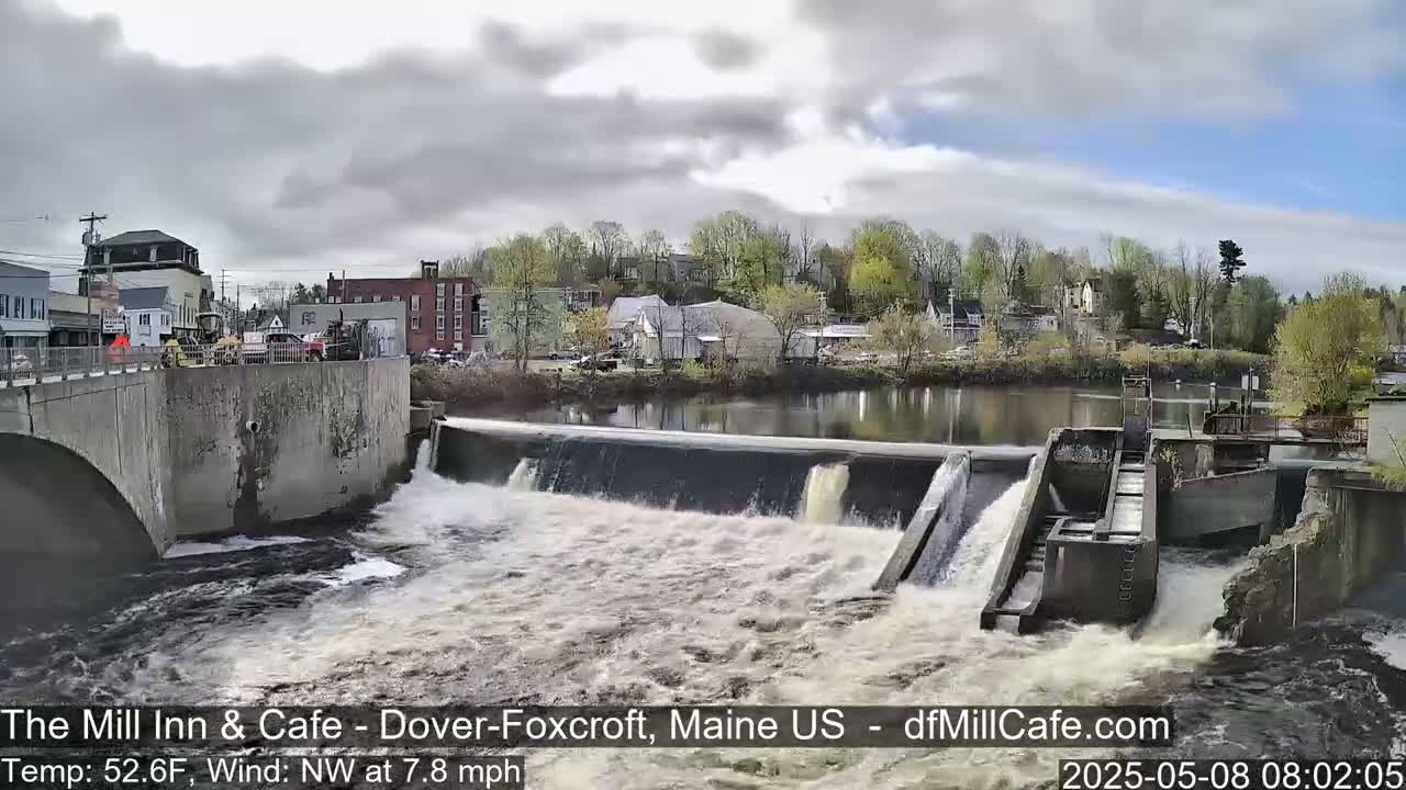 Water flows over a dam in a town under a partly cloudy sky.