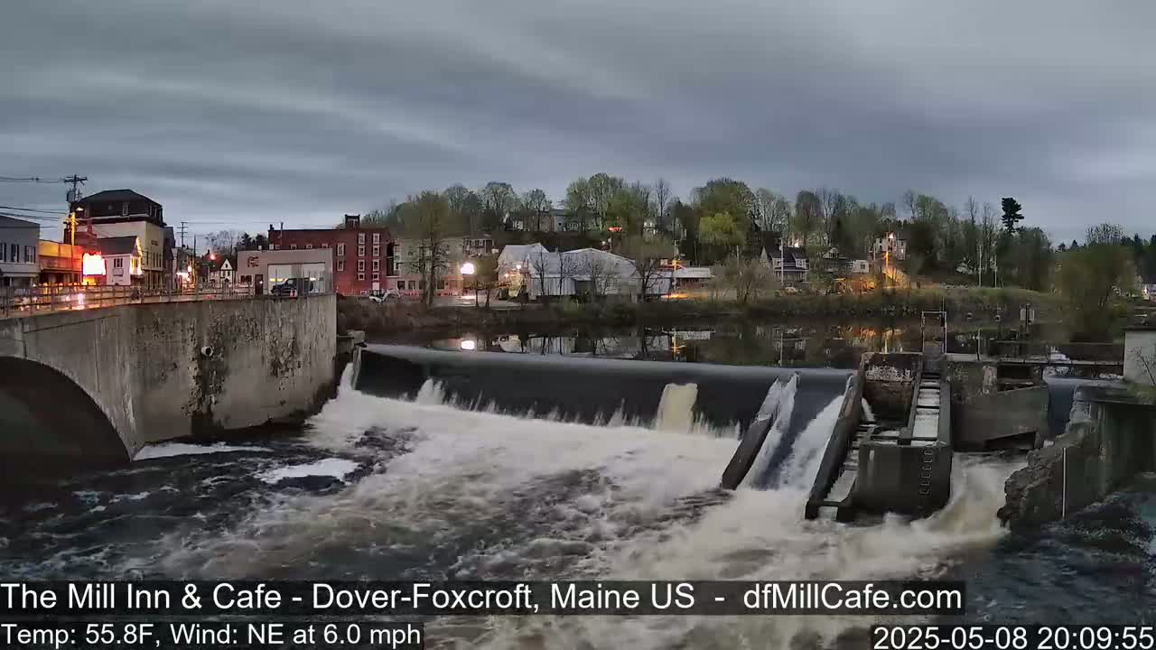 A river flows over a dam in a town under a cloudy evening sky.