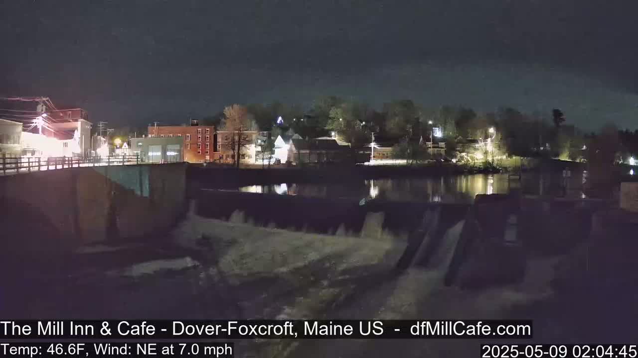 A nighttime view of a dam with water flowing over it, overlooking a town with illuminated buildings under a clear, dark sky.