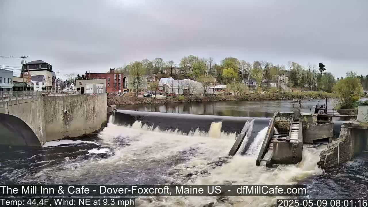 A river flows over a dam in a town under an overcast sky.