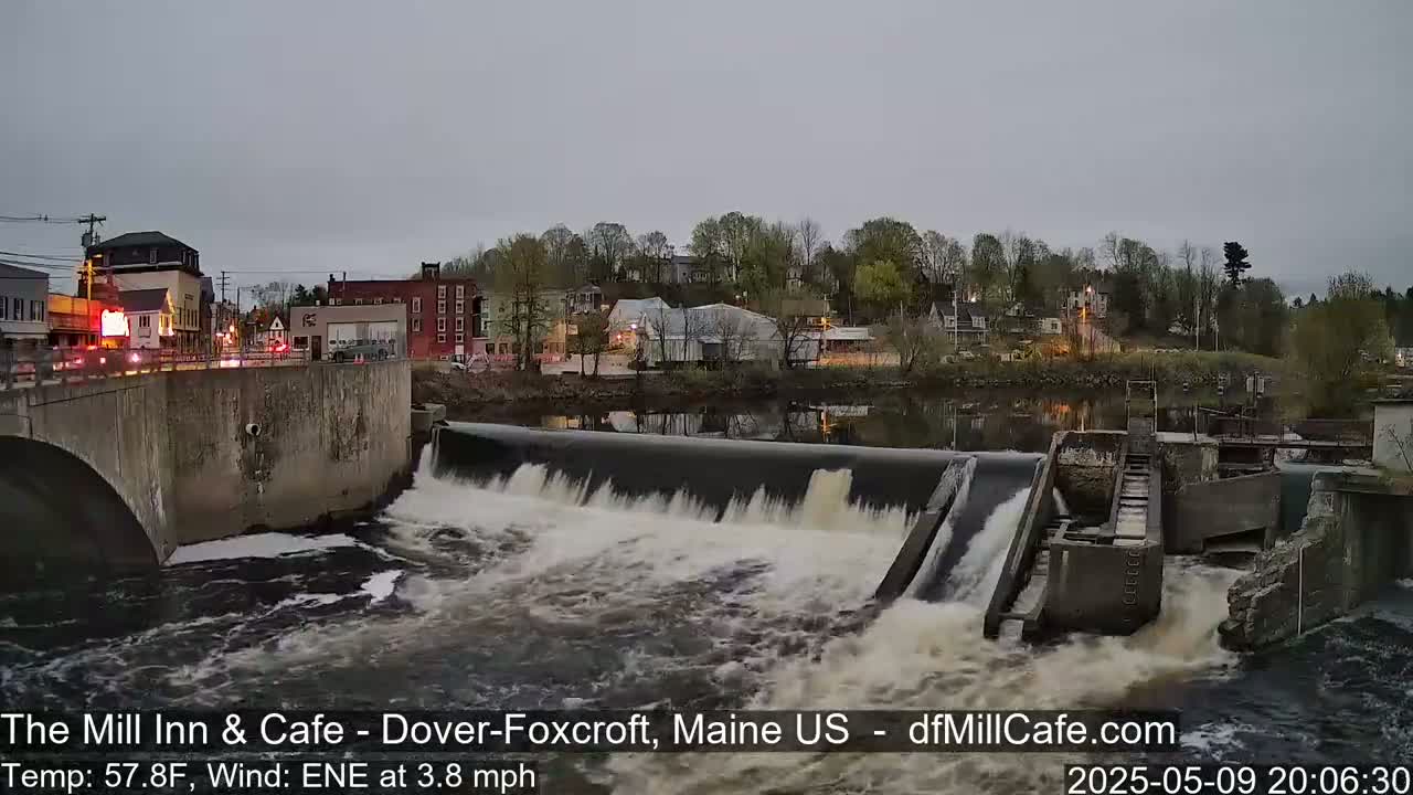 A river flows over a dam under an overcast sky, with a town visible in the background.
