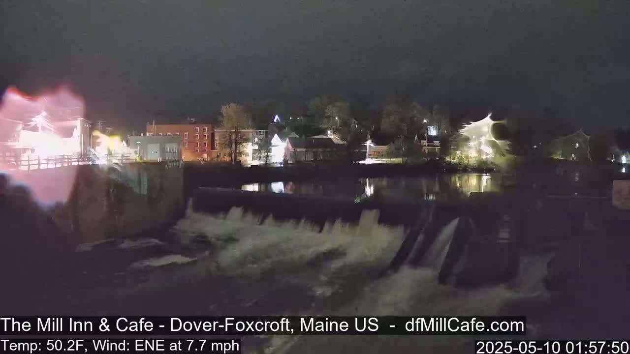 A nighttime view of a dam with water flowing over it, illuminated buildings across a calm river, and a somewhat hazy sky.