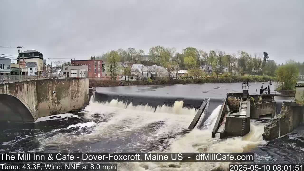A river flows over a dam in a town under an overcast sky.