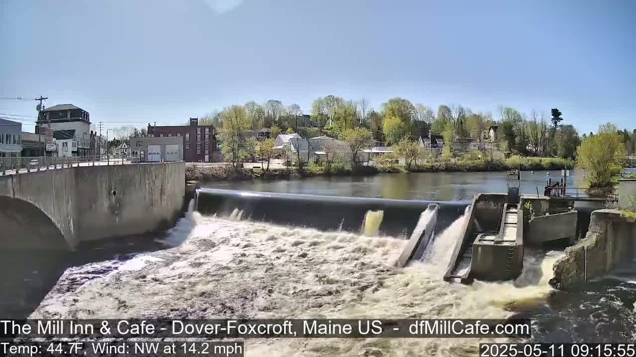 A dam releases water into a river flowing past a small town on a sunny day with a northwest wind.