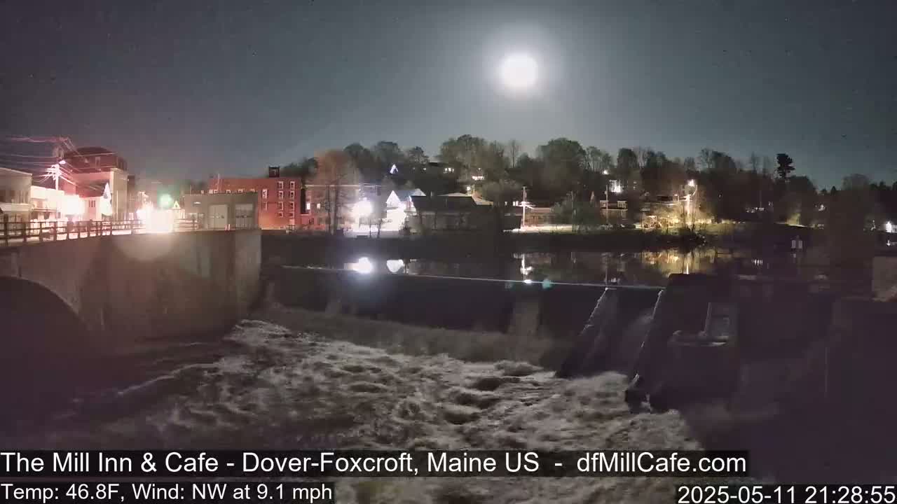 A moonlit night scene shows a river flowing over a dam past a small town with lights visible under a mostly clear sky.
