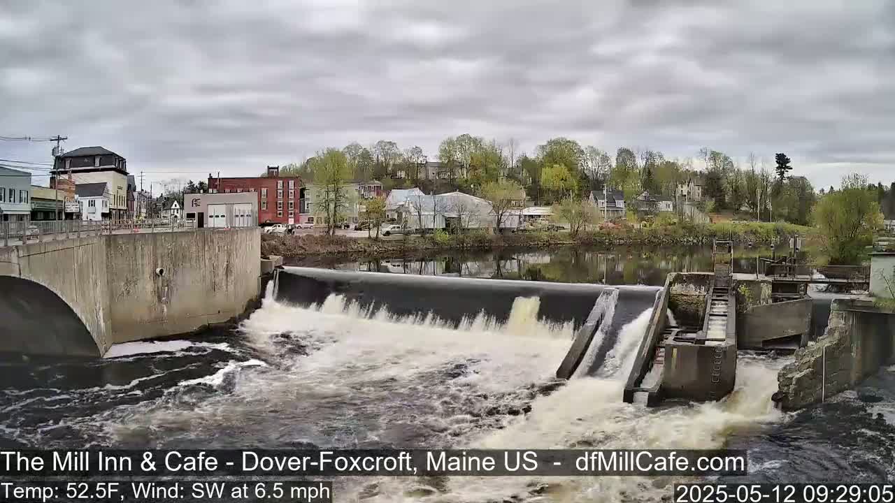 A river flows over a dam in a town under an overcast sky.