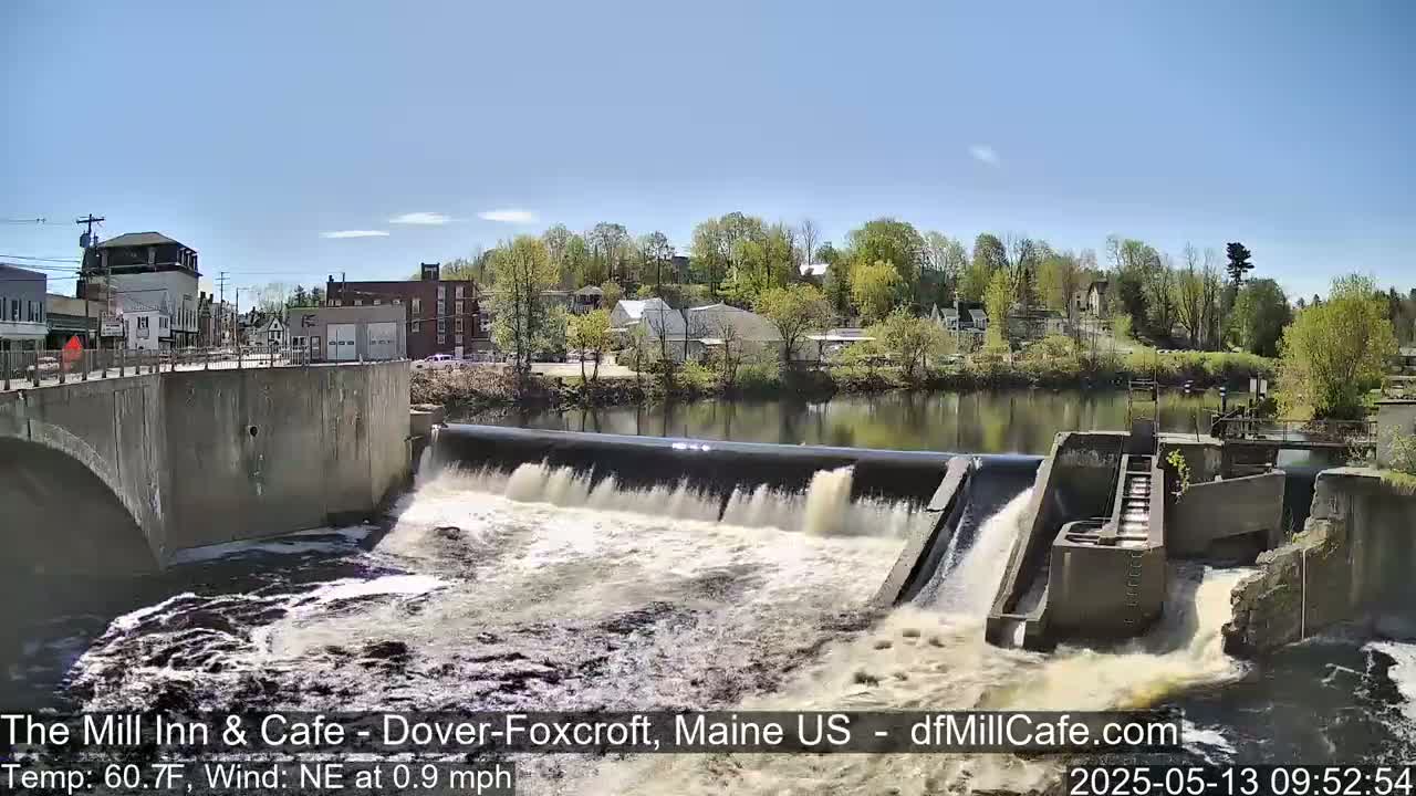 A dam with water flowing over it in a town on a sunny day.