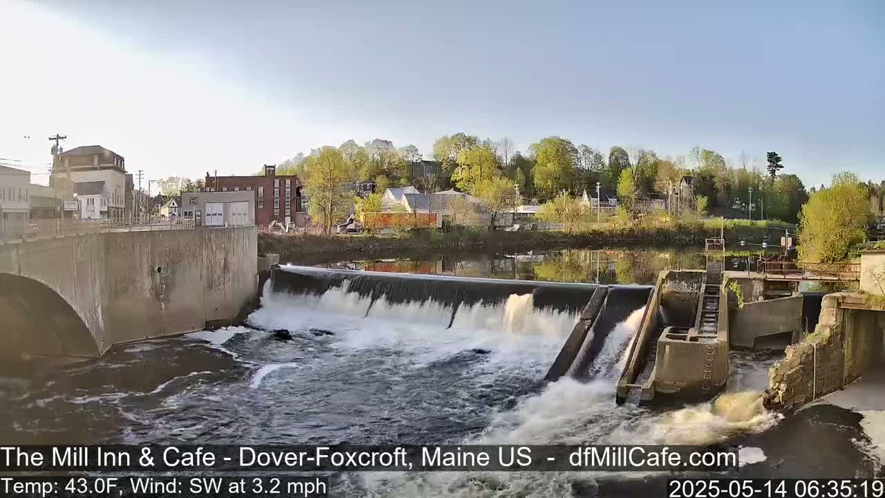 A river flows over a dam in front of a town under a clear, sunny sky.