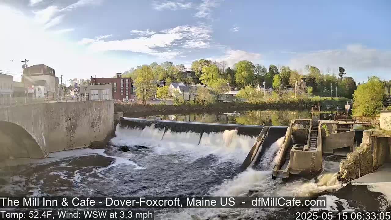 A dam with water flowing over it is situated in front of a town with buildings and lush green trees under a partly cloudy sky.