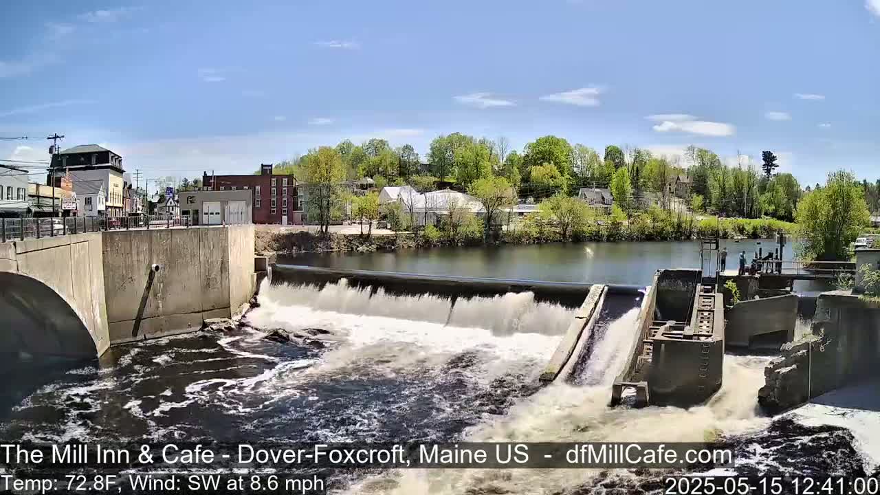 A dam releases water into a river flowing past a small town under a sunny sky.