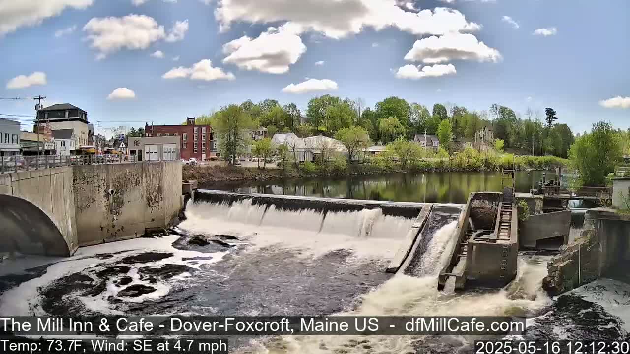 A river flows over a dam in a town on a partly cloudy day.