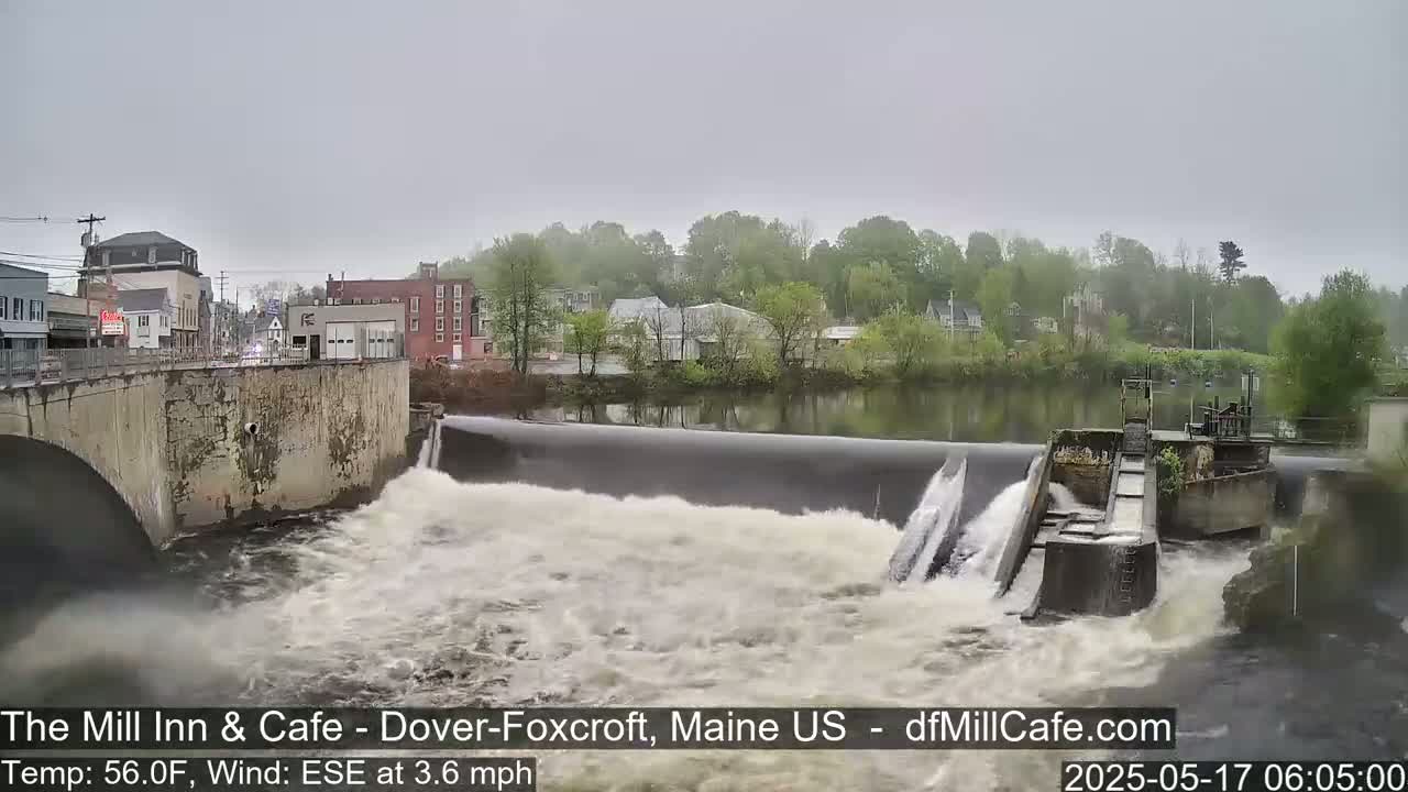 A dam with water rushing over it, a bridge, and buildings are visible under an overcast sky.