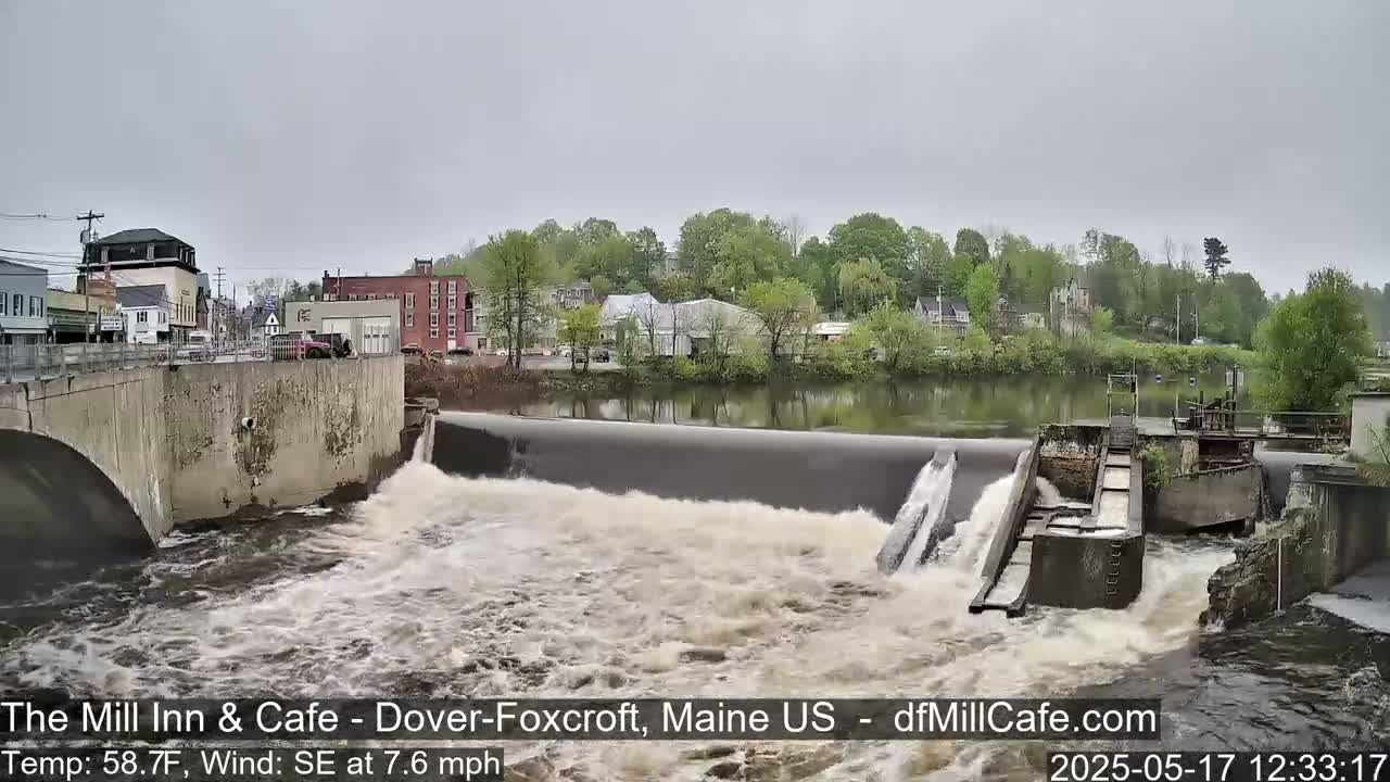 Water flows over a dam in front of a town, under an overcast sky.