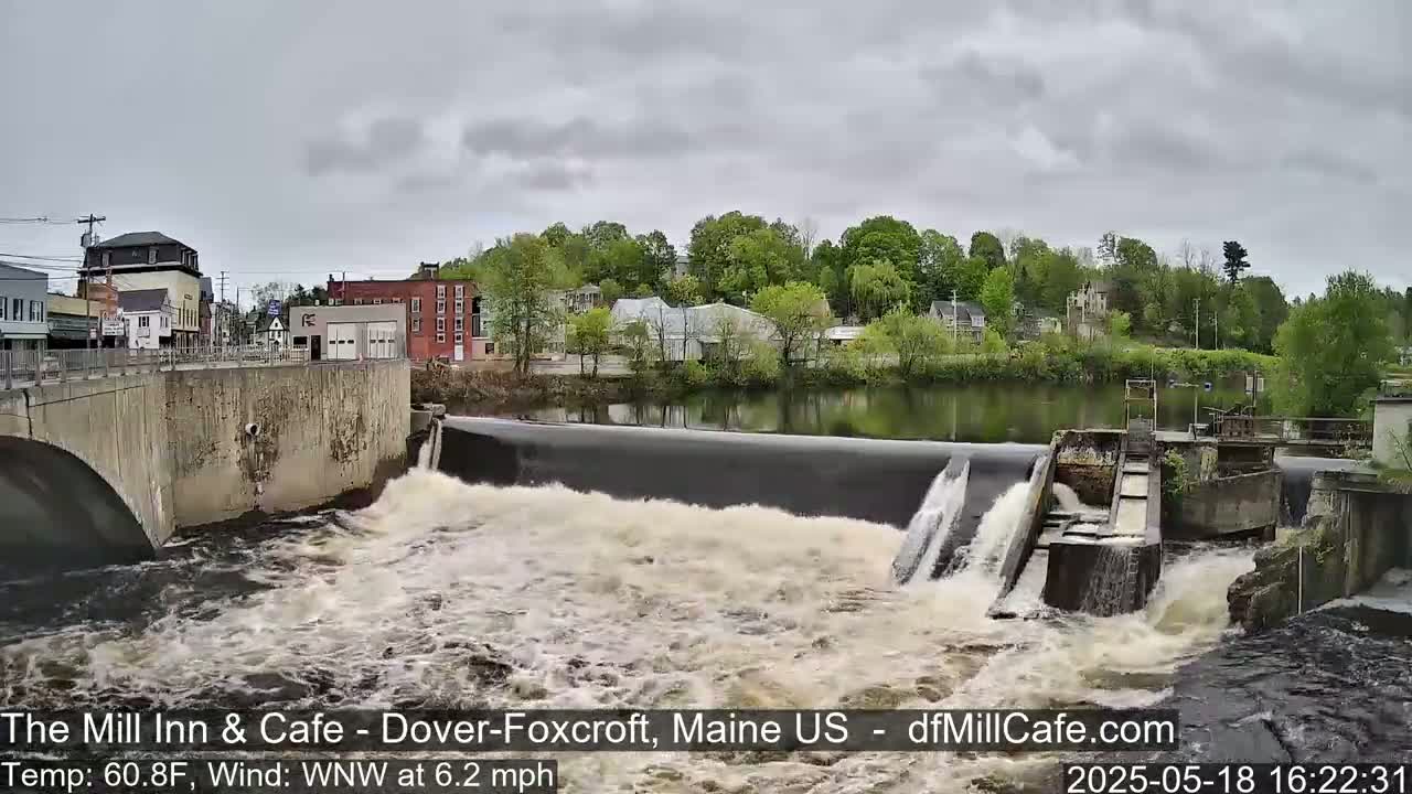 Water rushes over a dam in front of a small town under an overcast sky.