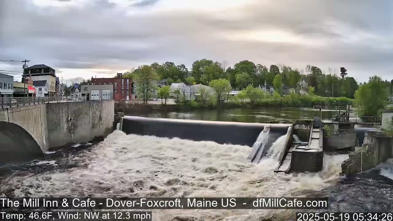 A dam releases water into a rushing river, with a town visible in the background under an overcast sky.