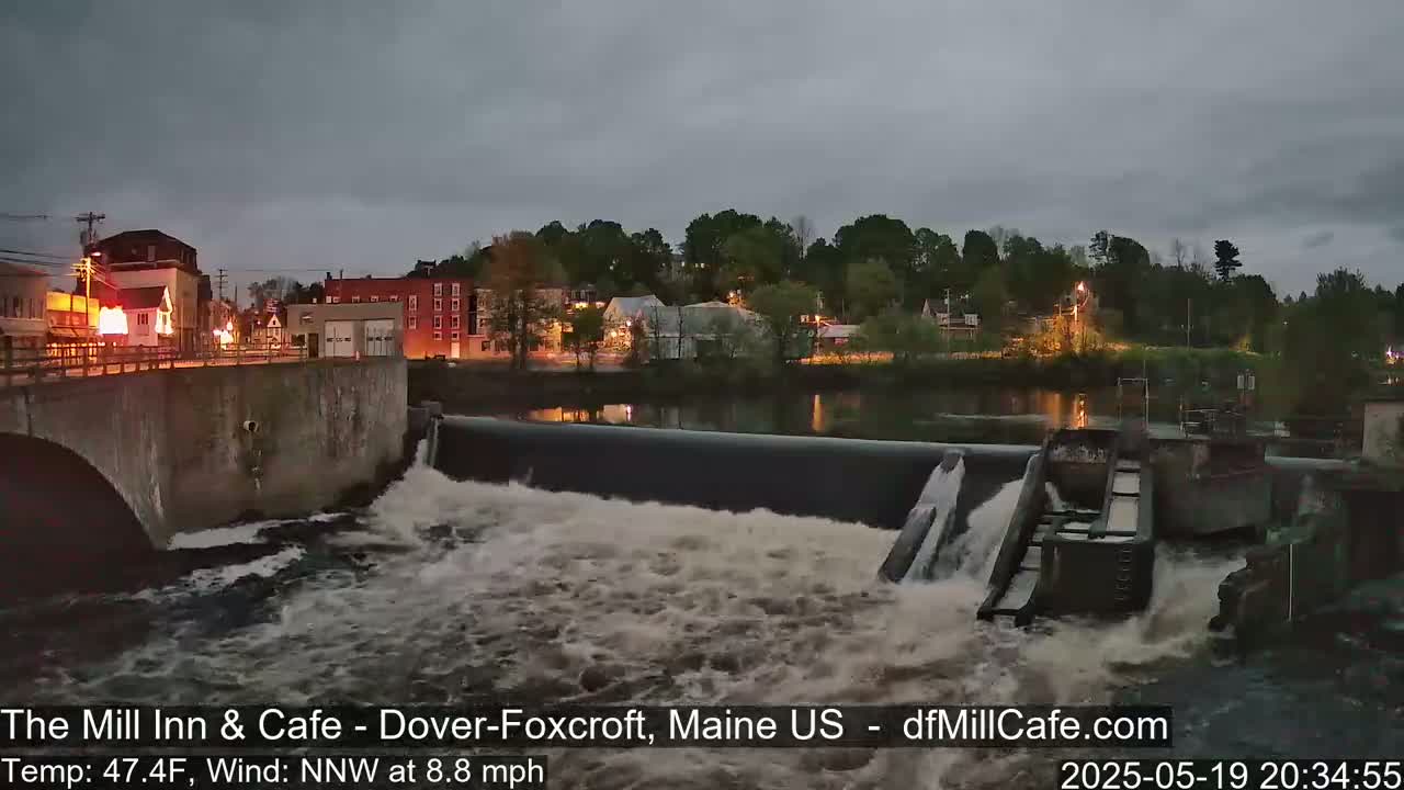 Water flows over a dam at dusk, past a town with lights on, under a cloudy sky.