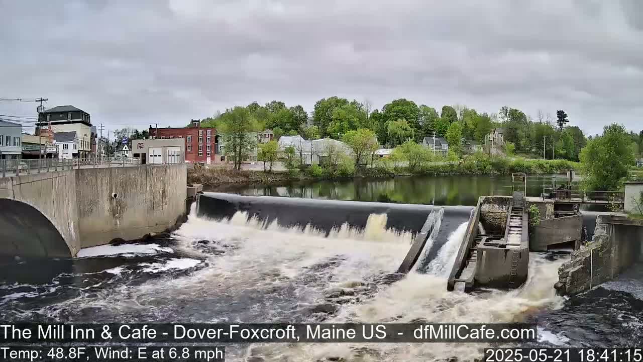 A river flows over a dam in a town under an overcast sky.