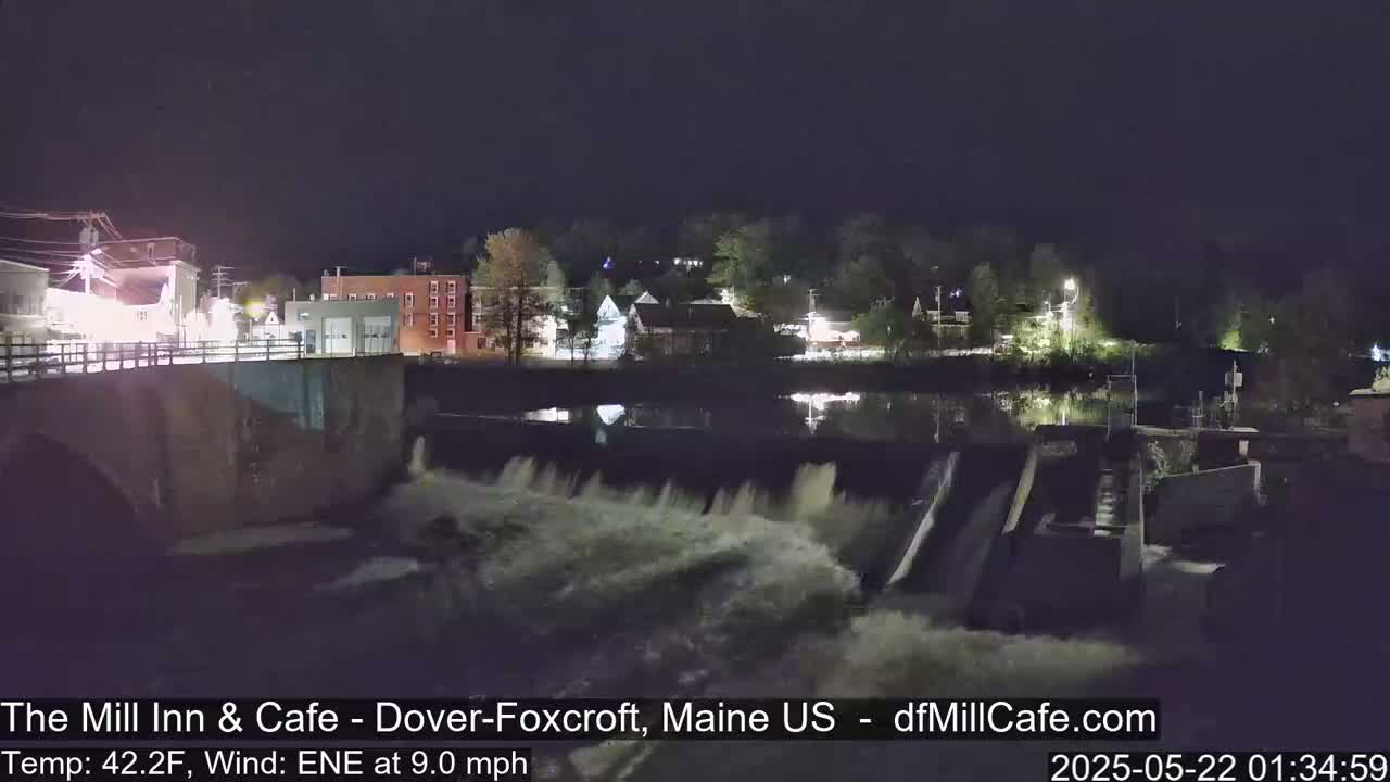A nighttime view of a dam with water cascading over it, illuminating a calm river reflecting the lights of a small town across the water under a clear sky.