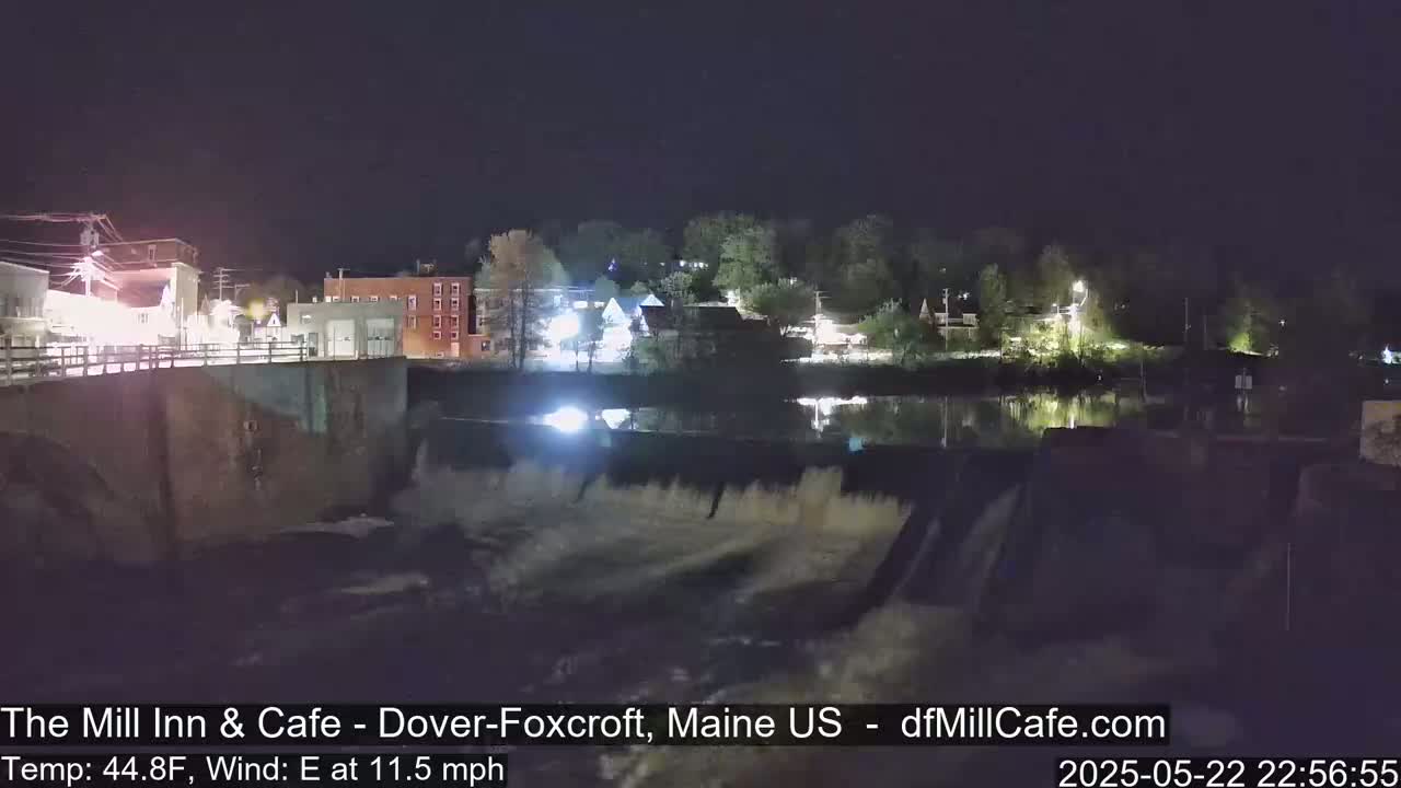 A nighttime view of a town's illuminated buildings and a dam with water flowing over it under a dark sky.