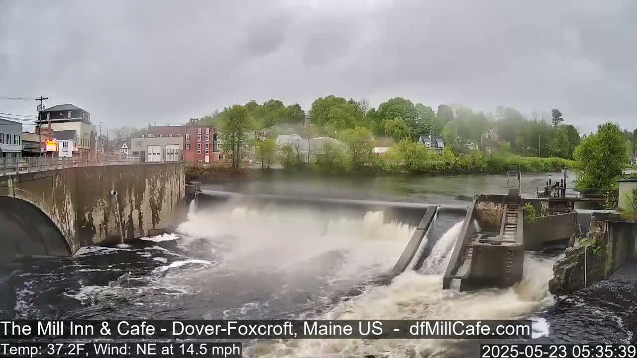 Water flows over a dam in a town, under an overcast sky with a northeast wind.
