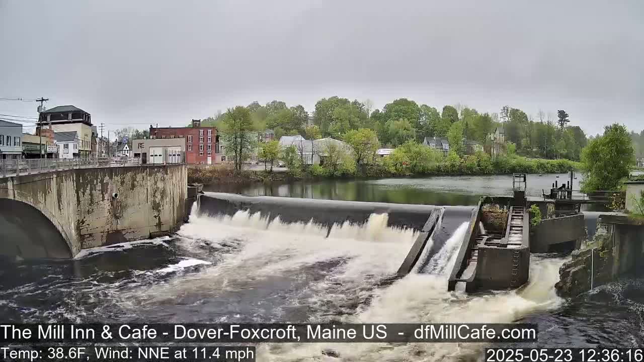 A dam with water rushing over it in a town under an overcast sky.