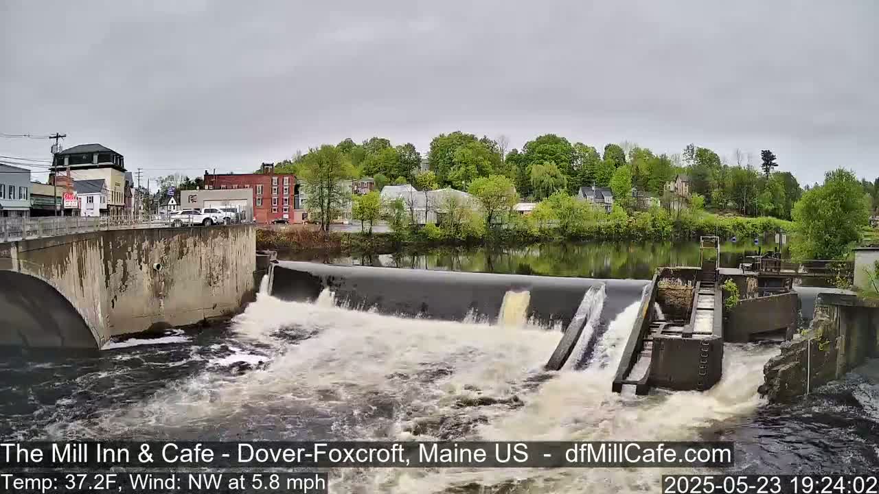 A dam with water flowing over it into a river, under an overcast sky, in front of a town with trees and buildings.
