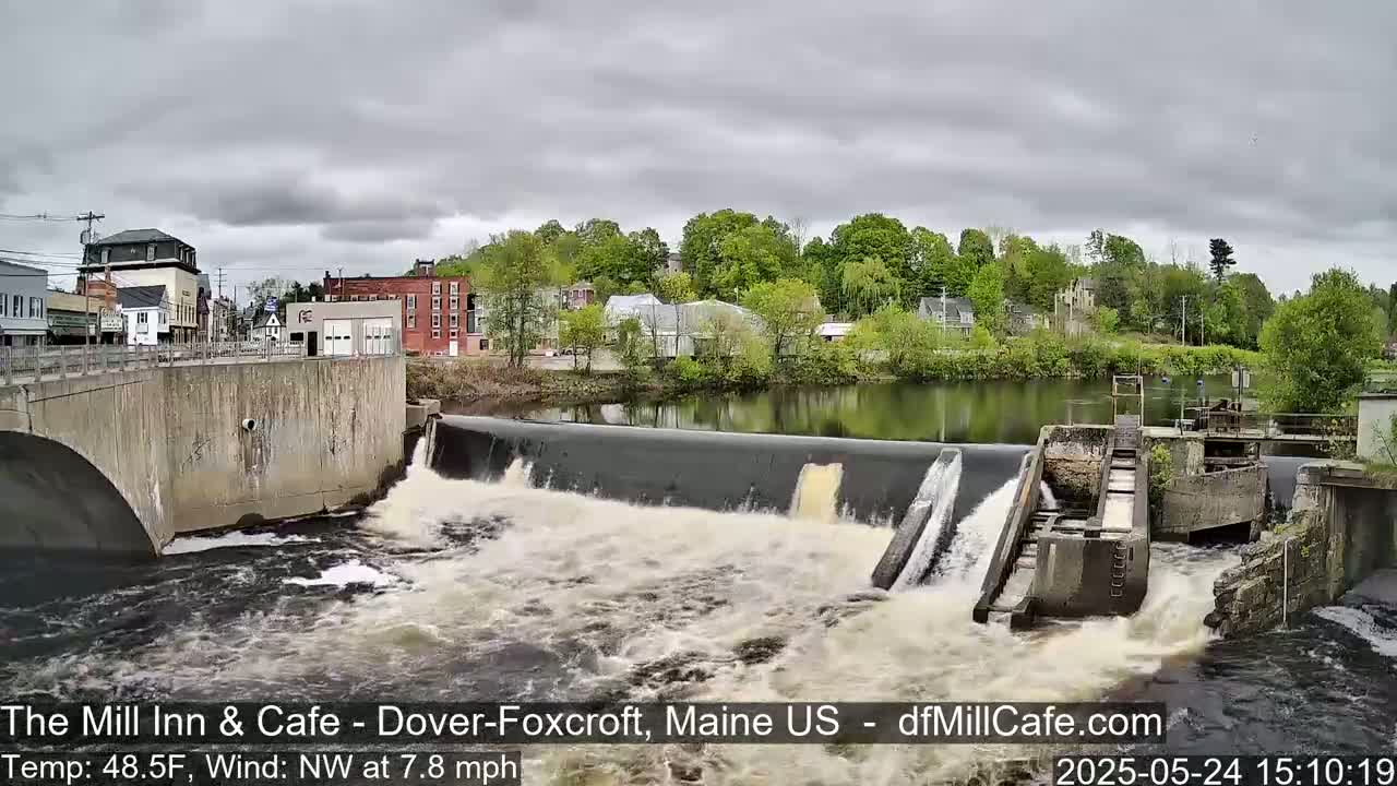 Water rushes over a dam in a town under a cloudy sky.