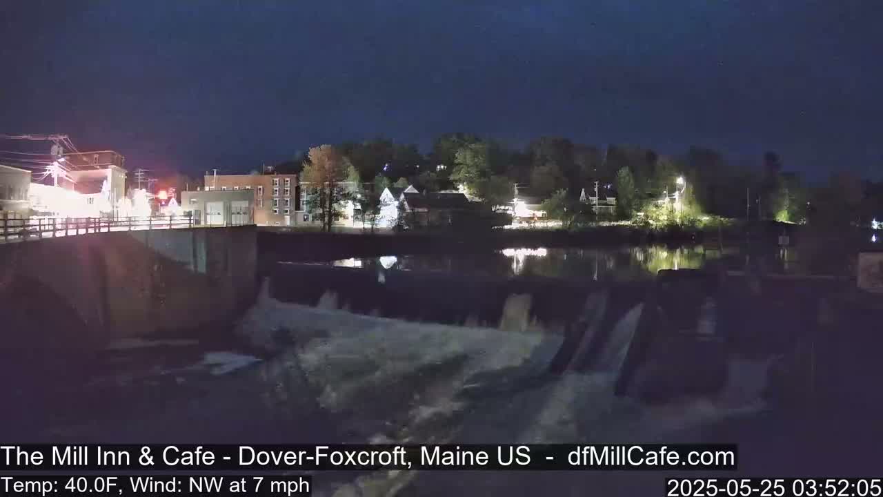A nighttime view of a dam with water flowing over it, overlooking a town illuminated by lights under a dark sky.
