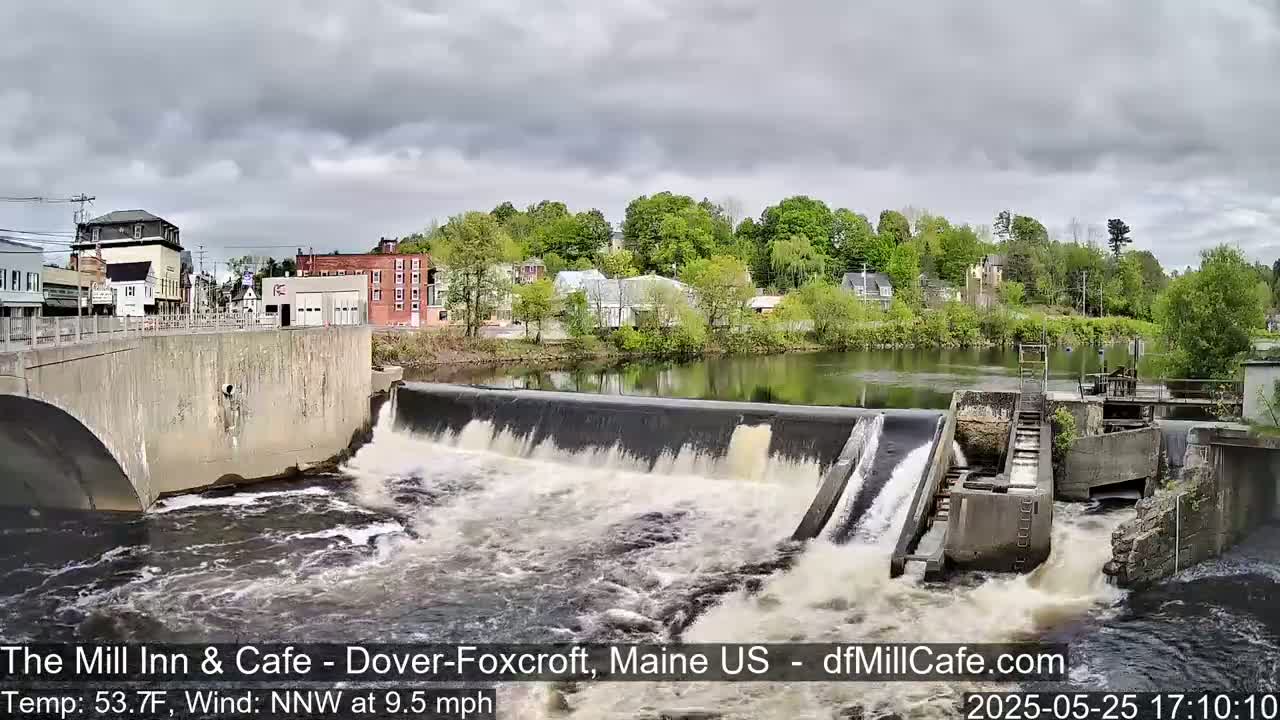 Water flows over a dam in a town, under a cloudy sky.