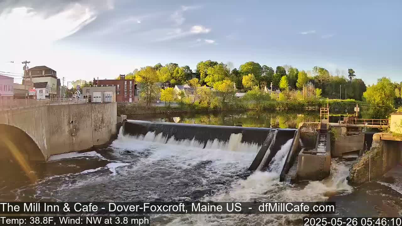 A dam with flowing water is situated in a town with buildings along a river under a partly cloudy sky.