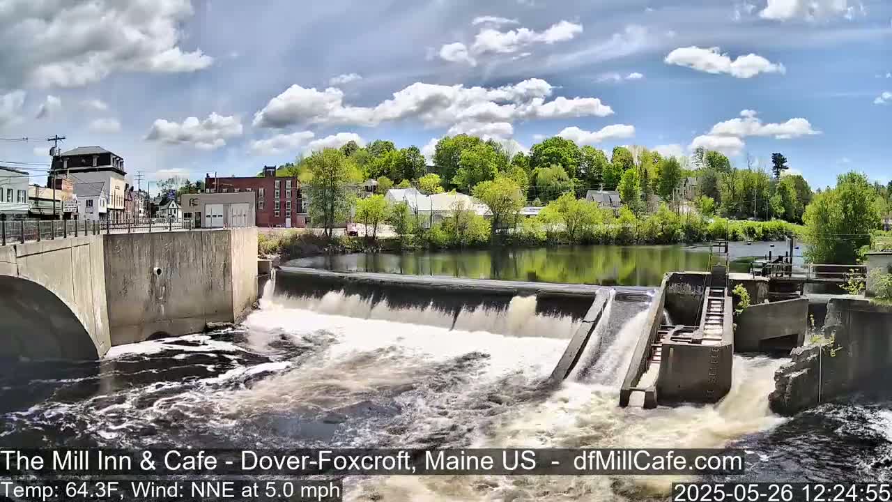 Water flows over a dam in a town under a partly cloudy sky.