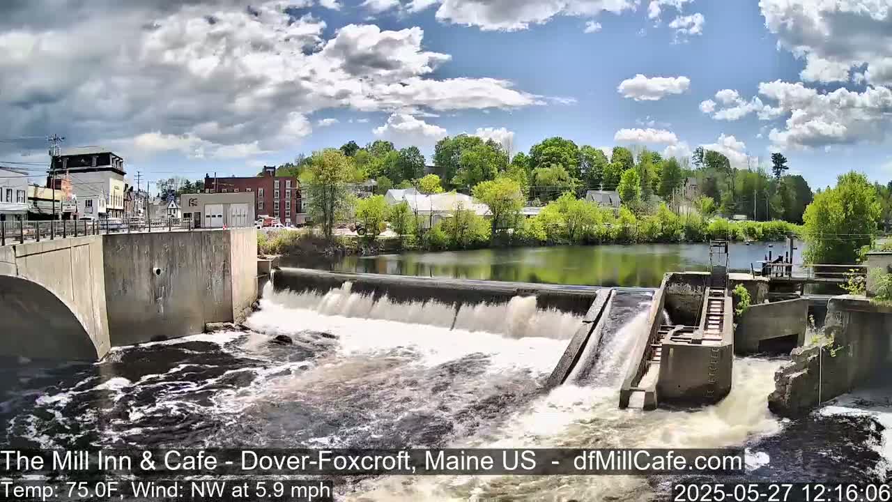 A dam with water flowing over it, situated in a town with buildings and lush green trees under a partly cloudy sky.