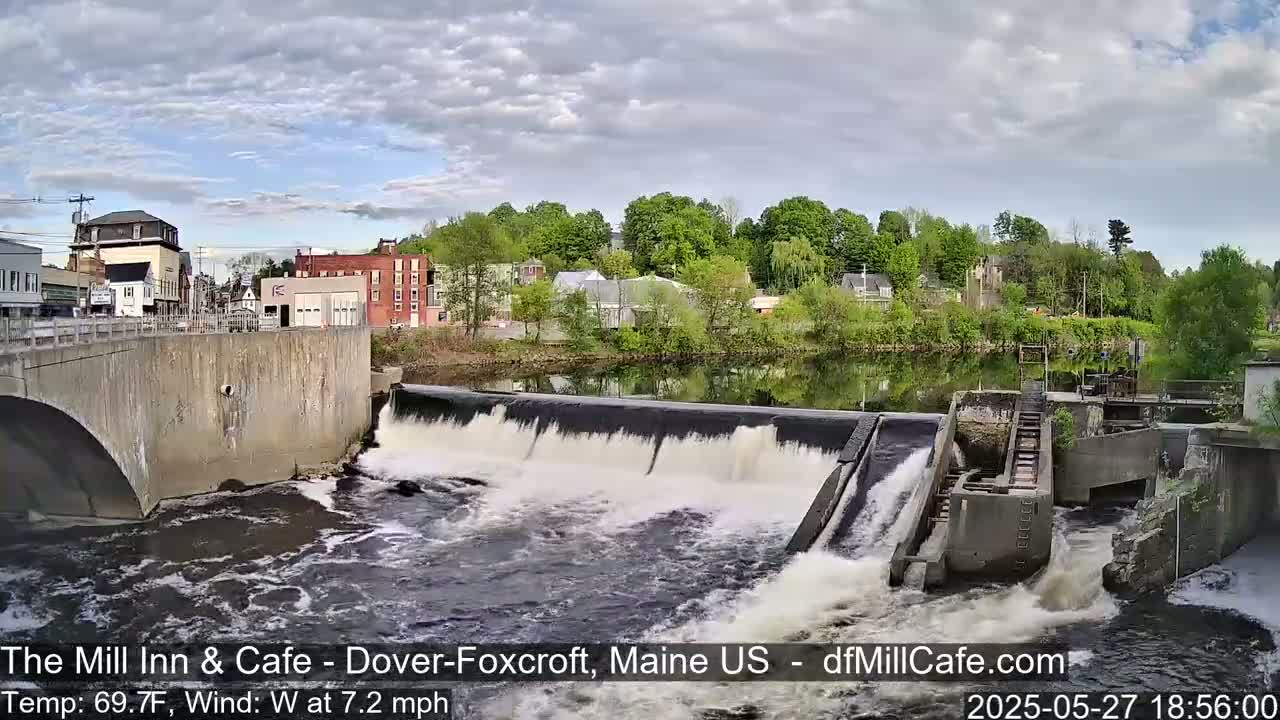 A dam with water flowing over it sits in front of a small town under a partly cloudy sky.