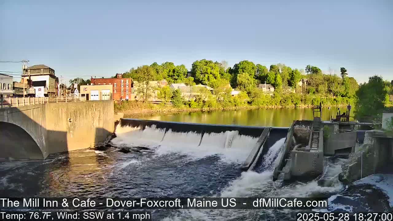 Water flows over a dam in a town, under a clear, sunny sky.