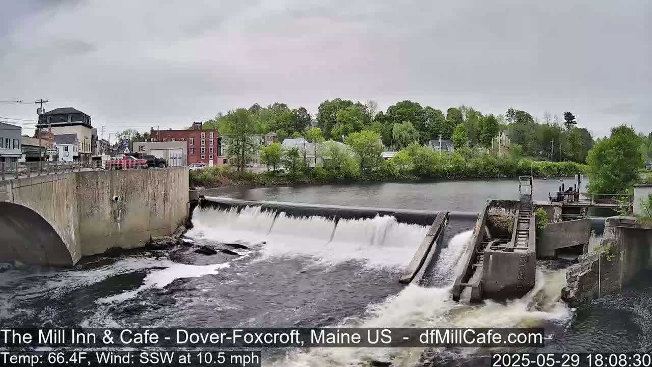 A river flows over a dam in a town under an overcast sky.