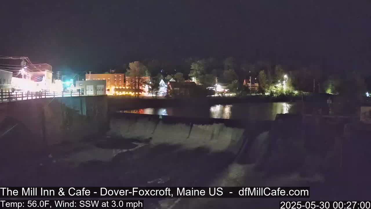 A nighttime view of a town's illuminated buildings and a dam with water flowing over it, under a clear sky.