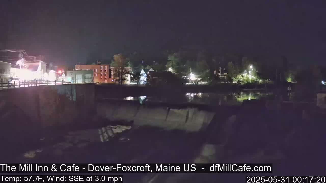 A nighttime view of a calm river reflecting lights from buildings across a dam, under a clear sky with a gentle south-southeast wind.