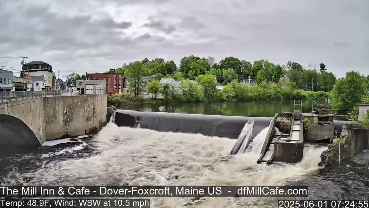 Water rushes over a dam in a town under an overcast sky.