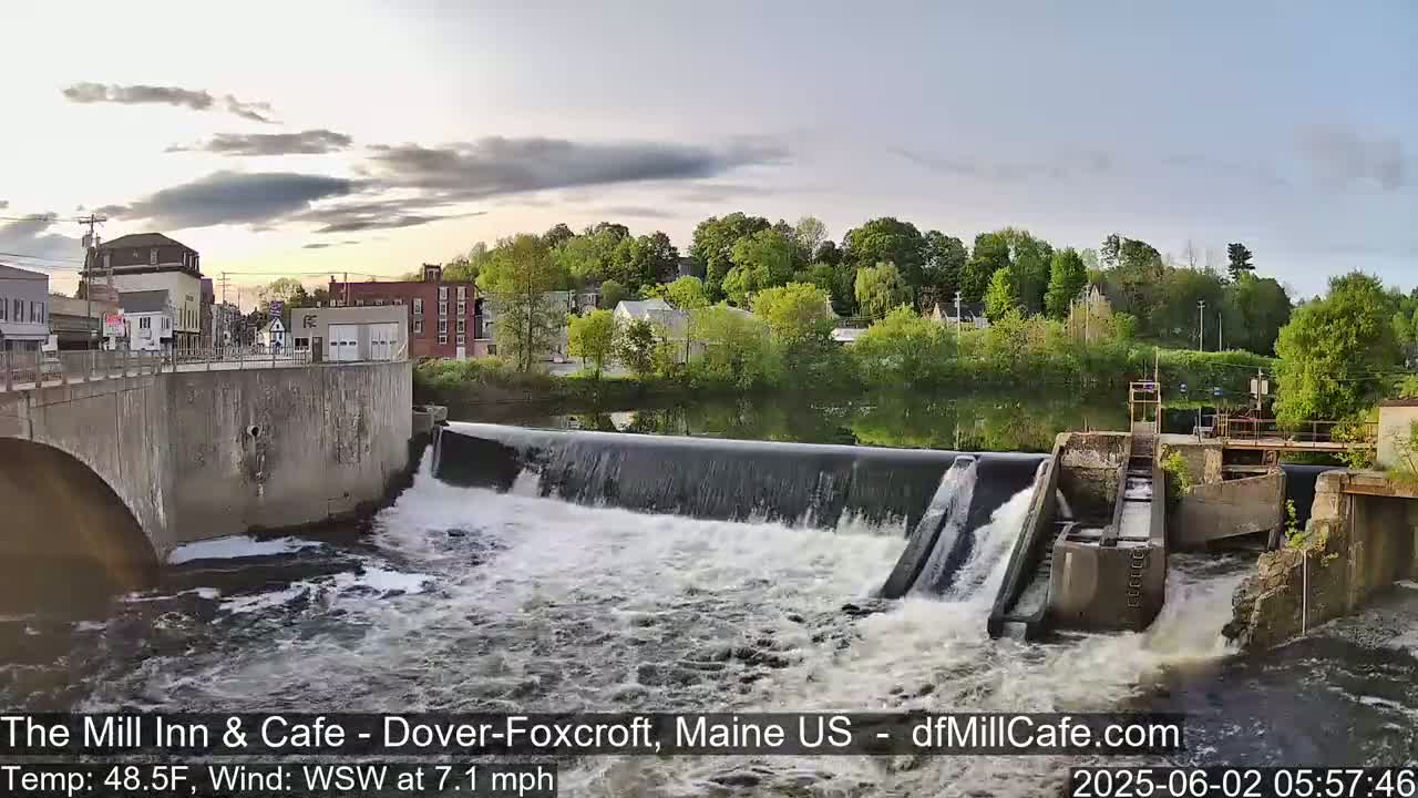 A river flows over a dam in a town under a partly cloudy sky.