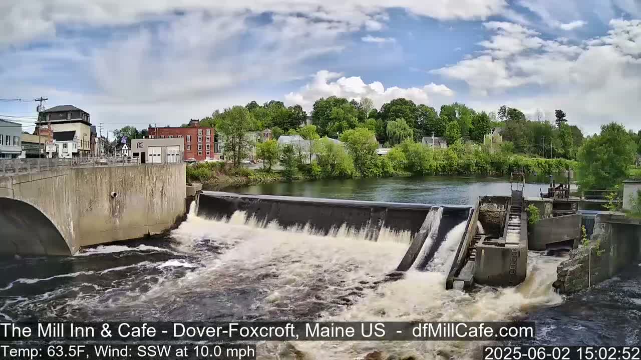 Water flows over a dam in a town beside a river under a partly cloudy sky.
