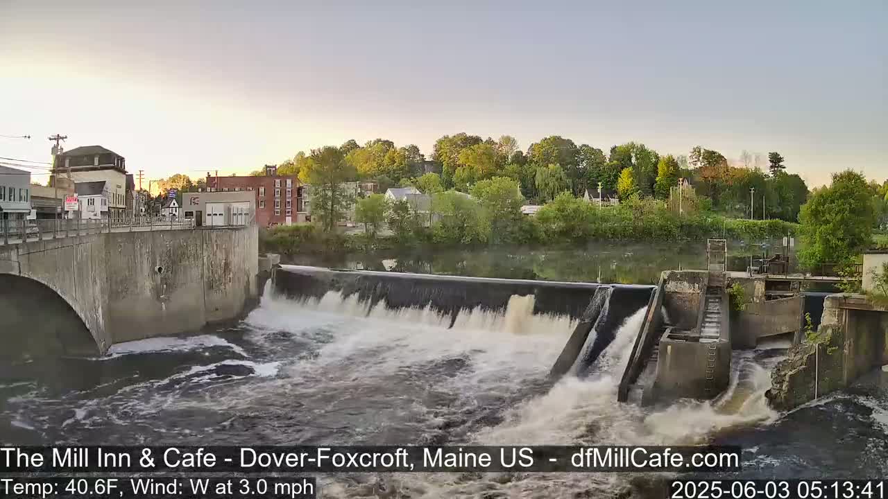 A dam with water flowing over it sits in front of a town at sunrise, under a mostly clear sky with a light westerly wind.