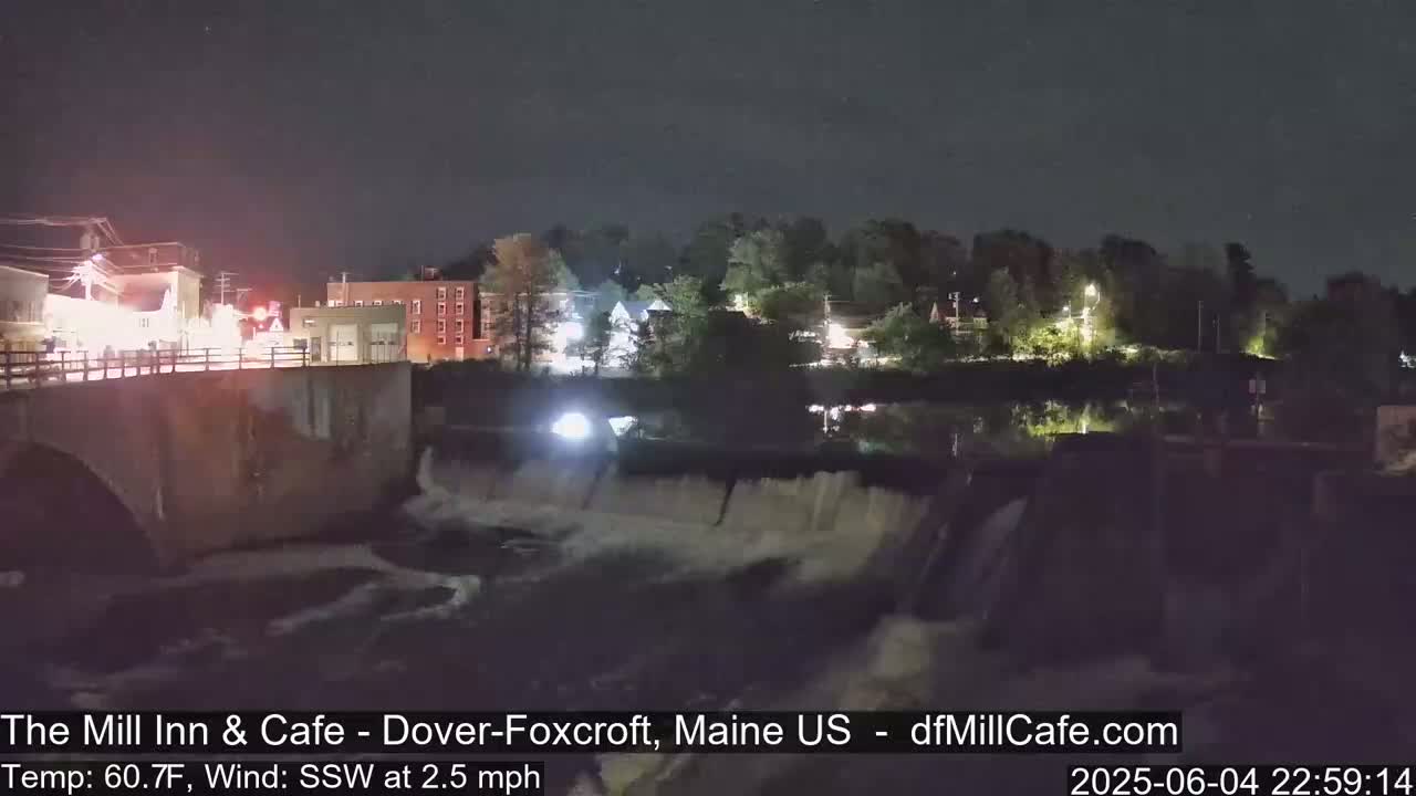 A nighttime view of a dam and river flowing through a town, under a clear sky with a slight southwest wind.