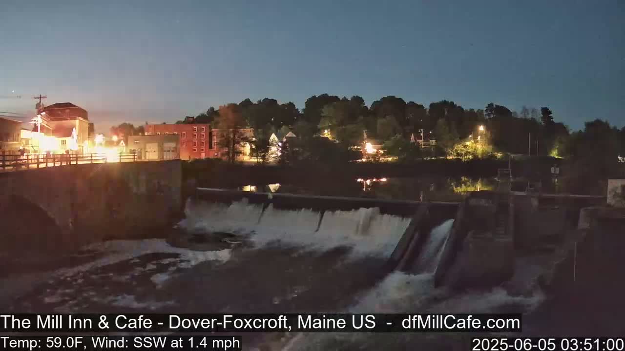 A nighttime view of a dam and river flowing over it, with a town's lights visible in the background under a twilight sky.